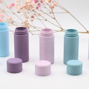 May include: A collection of colorful, textured bottles with matching screw-on lids. The bottles are in shades of blue, purple, and pink. The design features a diagonal, ridged pattern. A sprig of pink flowers is in the background.