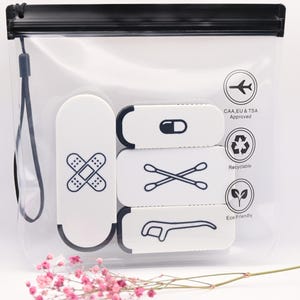 May include: A clear, rectangular plastic travel pouch with a black zipper and wrist strap. Inside are three white rectangular cases with black icons: a bandage, a pill, cotton swabs, and a dental flosser. Text includes CAA, EU & TSA Approved.