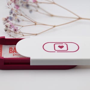 May include: A magenta and white sliding case with a rounded rectangular shape. The white lid features a magenta graphic of a pill capsule with a heart inside. Inside the case, a white bandage wrapper with red text is partially visible.