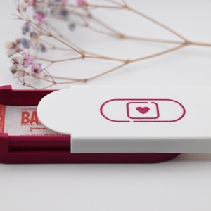 May include: A burgundy and white pill case with a band-aid design and a heart icon. The case is open, revealing a packet of medication. Delicate, dried flowers are in the background.