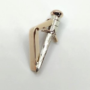 May include: A metallic tie bar featuring a two-tone design with a polished rose gold-colored angular clip attached to a silver-colored base shaped like a small sword or dagger with a rounded pommel at the top end.