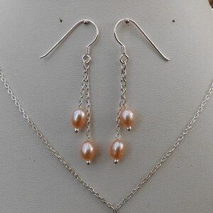May include: A pair of silver earrings with a delicate chain design and three pink pearls hanging from each earring. The earrings are displayed on a white background.