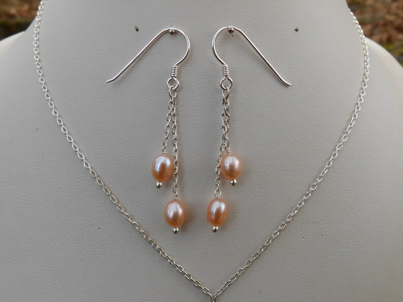 May include: A pair of silver earrings with a delicate chain design and three pink pearls hanging from each earring. The earrings are displayed on a white background.