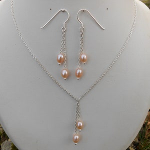 May include: A silver necklace and earrings set with a delicate chain and three pink pearls dangling from each piece.