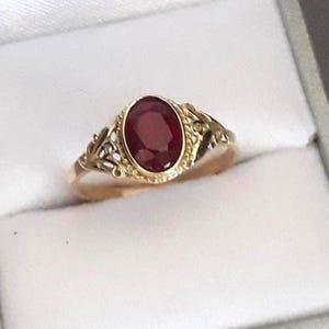 Può includere: Un anello in oro con una pietra preziosa rosso scuro di forma ovale, incastonata in una fascia decorativa in stile filigrana. La pietra centrale è circondata da una montatura in oro testurizzato e le spalle dell'anello presentano dettagli traforati.