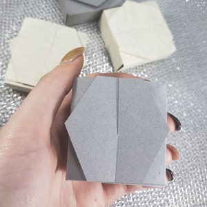 May include: Three gray origami boxes, one held in a hand. The boxes are folded into a square shape with a pointed top.