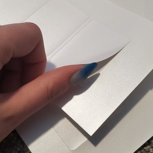 May include: A stack of white, rectangular, adhesive labels with one label being peeled back to reveal a silver, shimmery surface.