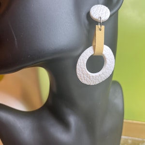 Peut inclure: Boucles d'oreilles pendantes blanches et brunes avec une finition texturée. Les boucles d'oreilles présentent un petit cercle blanc en haut et un cercle blanc plus grand avec une lanière en cuir marron au milieu.