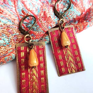 May include: Two rectangular earrings with a red and gold woven design. Each earring features a teardrop-shaped, light-brown bead suspended from a bronze-coloured metal frame. The earrings are shown on patterned fabric.