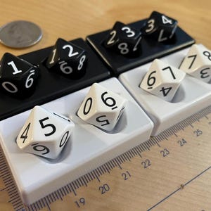 May include: Two black and two white rectangular trays, each holding two ten-sided dice. The black trays contain black dice with white numbers, and the white trays contain white dice with black numbers. A ruler shows measurements in centimeters.