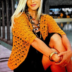 Peut inclure: Une femme portant un cardigan en crochet orange avec une robe noire et un collier à plusieurs rangs. Elle est assise sur une surface en bois avec un arrière-plan flou.