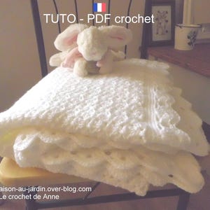 Peut inclure: Une couverture bébé en crochet blanc avec une bordure en dentelle délicate. La couverture est pliée et exposée sur une chaise en bois avec un petit jouet de lapin blanc sur le dessus. Le texte "TUTO - PDF crochet" est visible sur l'image.