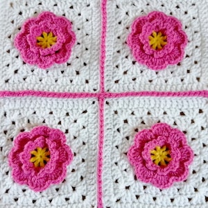 Peut inclure: Une couverture en crochet blanche avec quatre motifs de fleurs roses au centre de chaque carré.