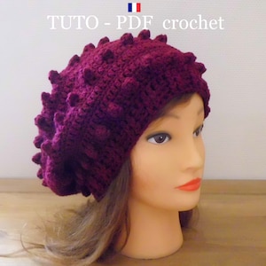 Peut inclure: Un béret en crochet de couleur bordeaux avec un design texturé et bosselé. Le béret est sur une tête de mannequin avec des cheveux bruns.