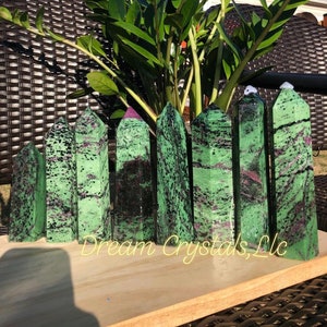 May include: A collection of eight green and black gemstone points with a pink hue. The points are arranged in a row on a wooden surface. The text "Dream Crystals, LLC" is visible in the image.