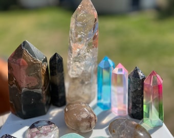 May include: A collection of various crystals and stones, including clear quartz, black obsidian, pink amethyst, and green aventurine. The crystals are arranged on a white surface with a green heart-shaped dish.