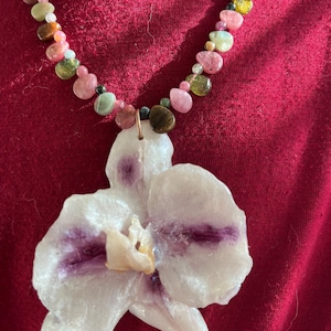 Peut inclure: Un collier avec un pendentif en forme d'orchidée blanche aux accents violets et une chaîne de perles de pierres précieuses colorées. Le collier est présenté sur un fond rouge, mettant en valeur le design floral et les perles vibrantes.