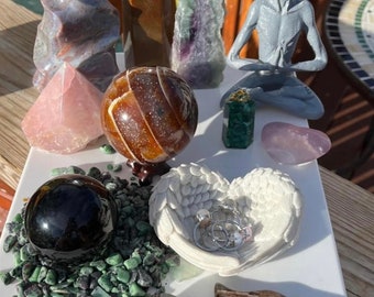 May include: A collection of crystals, stones, and other objects arranged on a white surface. The objects include a grey alien figurine, a heart-shaped dish with wings, a black sphere, a brown sphere, a pink pyramid, a green crystal, a brown and white striped stone, and a brown and white striped stone.