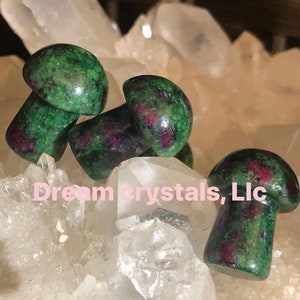 May include: Four green and pink gemstone mushroom shaped carvings on a bed of white crystal rocks. The text "Dreamcrystals, Llc" is visible in the image.
