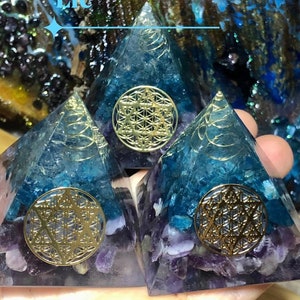 May include: Three small pyramids made of clear resin with embedded amethyst and blue crystals. Each pyramid has a gold metallic flower of life symbol on the front.