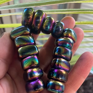 Puede incluir: Una pulsera iridiscente hecha de cuentas rectangulares. Las cuentas cambian de color, mostrando tonos de verde, morado y dorado. La pulsera se sostiene en una mano, con un fondo borroso de hojas verdes.