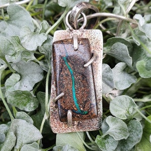 May include: A silver pendant necklace featuring a rectangular, dark brown stone with a vibrant teal vein. The stone is set in a textured silver frame, with silver accents. The necklace is displayed on a bed of green foliage.