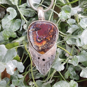 May include: A silver pendant necklace featuring a large, oval-shaped opal stone with a unique pattern of red, brown, and yellow hues. The pendant is suspended from a silver chain and has a decorative silver backing with a patterned design.