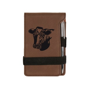 May include: Brown leatherette notepad with a black cow head graphic. The notepad has a black elastic band and a silver pen holder with a silver pen. The notepad is rectangular and has a smooth texture.