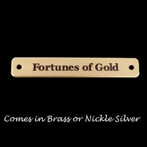 May include: A gold-colored metal tag with the text "Fortunes of Gold" engraved on it. The tag has two holes on either side. The text below the tag reads "Comes in Brass or Nickel Silver".