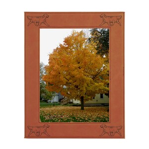 May include: A brown leather picture frame with a cutout of a golden retriever dog in each corner. The frame is holding a photo of a yellow tree in the fall with leaves on the ground.