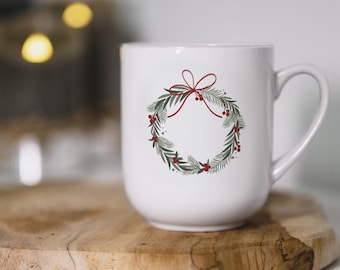 May include: White ceramic mug with a red bow and green wreath design. The wreath is decorated with red berries.