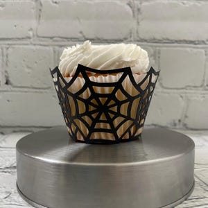 May include: A cupcake with white frosting sits in a black spiderweb-design cupcake wrapper. The cupcake is on a silver metal cake stand, set against a white brick wall background. A festive treat for Halloween.