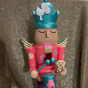 Könnte beinhalten: Ein hölzerner Nussknacker mit einem rosa-roten Outfit, einer türkisfarbenen Krone mit einem weißen Vogel und einem rosa Ei und goldenen Flügeln. Der Nussknacker hält eine Trommel mit einem rosa Bären und einen Schlagstock mit einem Löwen.