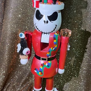 May include: A decorative wooden nutcracker in the style of Jack Skellington from The Nightmare Before Christmas. The figure is painted in red, white, and black, with a hat featuring Halloween-themed decorations. It stands on a black base with additional Halloween accents.