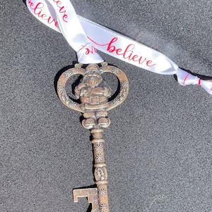 Könnte beinhalten: Dekorativer Schlüsselanhänger mit einer Santa Claus-Figur. Der Schlüssel ist metallisch mit Glitzer und Schneeflocken-Details. Ein weißes Band mit dem Wort "believe" in roter Schrift ist am Schlüssel befestigt.