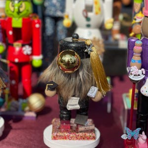 May include: A whimsical, handcrafted nutcracker figurine. The figure is dressed in a black graduation gown and cap, with a long, brown beard. It stands on a white base with books and glitter. A gold ornament and tassel add detail.