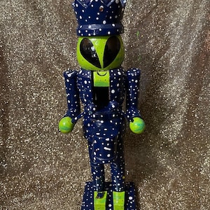May include: A whimsical wooden nutcracker in the shape of an alien. The figure is painted in navy blue with white polka dots, a green head, and a crown. The alien has black eyes and green hands and feet. The nutcracker stands on a square base.