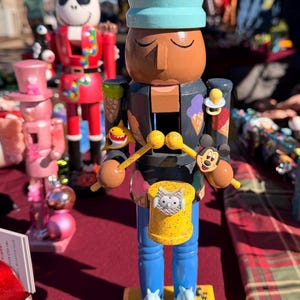 May include: A wooden nutcracker figure wearing a light blue crown with an orange fish accent. The dark gray torso features ice cream cone graphics, a pacifier, and a yellow drum with an owl. It has blue legs, white shoes, and a yellow base with puzzle pieces.