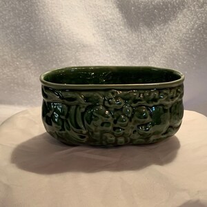 May include: A green ceramic planter with an embossed design of grapes and leaves. The planter is oval shaped and has a smooth, glazed finish.