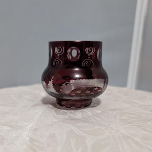 May include: A small, dark red glass vase with a white etched design. The vase has a round body and a flared top.