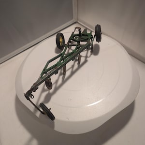May include: A vintage green metal toy farm plow implement featuring a long frame, multiple circular metal discs, and four small black rubber-tired wheels. The agricultural equipment model has a hitch attachment point and weathered paint finish.