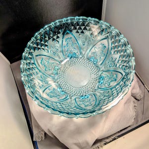 May include: A clear, light blue glass bowl with a decorative floral pattern. The bowl has a diamond-cut design around the rim and floral accents. The bowl is sitting on a light grey cloth.