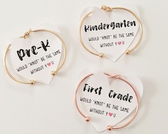 Pode incluir: Três pulseiras de ouro e ouro rosa com etiquetas em forma de coração. As etiquetas têm o texto "Pre-K", "Kindergarten" e "First Grade" com a frase "Would "knot" be the same without you" em cada etiqueta.
