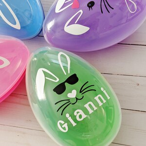 May include: Three plastic Easter eggs with bunny faces. The eggs are purple, blue, and green. The green egg has a bunny face with sunglasses and the name "Gianni" written on it. The purple egg has a bunny face with a pink bow and the name "Alex" written on it.