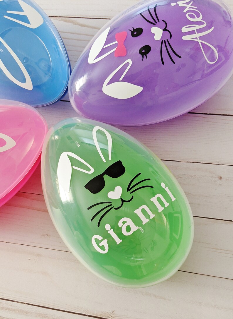 May include: Three plastic Easter eggs with bunny faces. The eggs are purple, blue, and green. The green egg has a bunny face with sunglasses and the name "Gianni" written on it. The purple egg has a bunny face with a pink bow and the name "Alex" written on it.