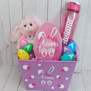 May include: A purple Easter basket with white polka dots and bunny ears. The basket contains a pink plush bunny, a pink plastic egg with the name "Ariana" on it, a clear water bottle with the name "Ariana" on it, and several plastic Easter eggs with the letter "A" on them.