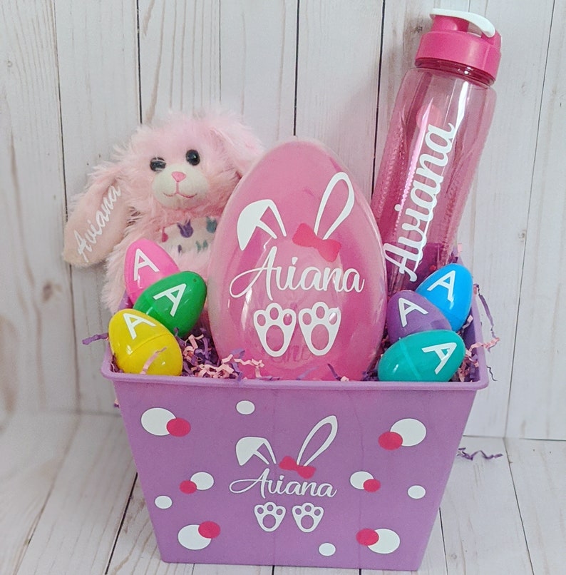May include: A purple Easter basket with white polka dots and bunny ears. The basket contains a pink plush bunny, a pink plastic egg with the name "Ariana" on it, a clear water bottle with the name "Ariana" on it, and several plastic Easter eggs with the letter "A" on them.