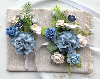 Puede incluir: Dos boutonnières de flores de papel azul y blanco con cinta blanca. Las boutonnières están hechas de flores de papel y vegetación.