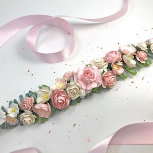 May include: A pink satin ribbon with a floral garland made of pink and white paper flowers and green leaves.