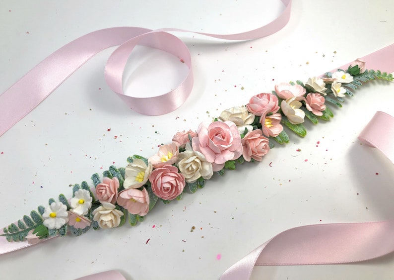 May include: A pink satin ribbon with a floral garland made of pink and white paper flowers and green leaves.